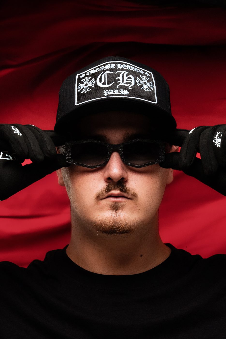 ALVA DJ in a Chrome Hearts cap and dark sunglasses against a red backdrop